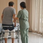 Patient vu de dos, assis au bord d’un lit d’hôpital, les deux genoux bandés après une prothèse, avec une kinésithérapeute qui l’aide à se lever près d’une fenêtre lumineuse